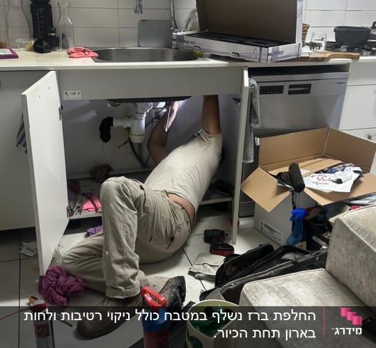 אדם מתקן צנרת מתחת לכיור במטבח עם כלים שונים סביבו